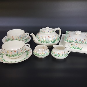 May include: A vintage tea set with a floral pattern. The set includes a teapot, two cups and saucers, a creamer, and a sugar bowl with a lid. The set is decorated with a pattern of green grass and colourful flowers.