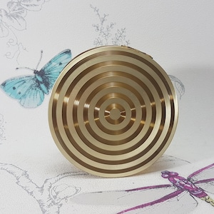 Op de afbeelding: Een goudkleurig, cirkelvormig object met concentrische ringen. Het object is gecentreerd, tegen een lichte achtergrond met een vlinder en bloemenmotief. Het ontwerp van het object doet denken aan een doel.