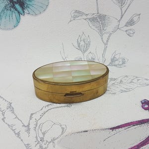 Könnte beinhalten: Ovale, antike Pillendose aus Messing mit einem karierten Deckel aus Perlmutt. Die Dose befindet sich vor einem weißen Hintergrund mit einem Blumen- und Libellenmuster. Die Dose ist geschlossen und hat einen kleinen Verschluss.