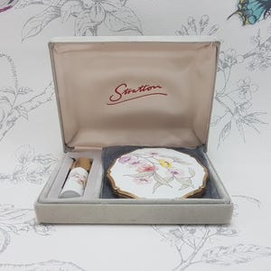 May include: A vintage Stratton compact set in a grey presentation box. The set includes a round white compact with a floral design and a matching small cylindrical bottle. The interior lid features the red Stratton logo on a pale satin lining.