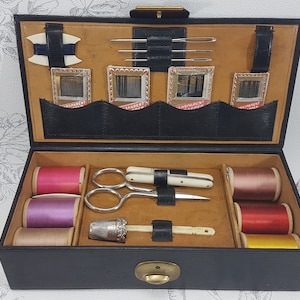 May include: A black leather sewing kit with a brown interior. The kit contains spools of thread in pink, purple, beige, red, and yellow. It also includes a thimble, a needle threader, a pair of scissors, and a small knife.