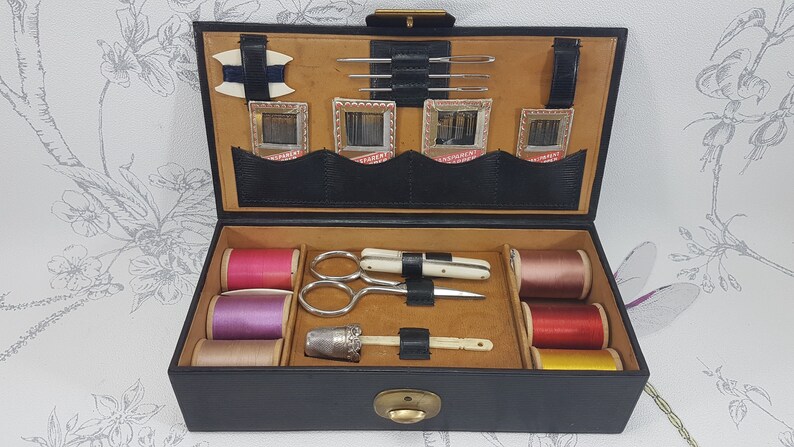 May include: A black leather sewing kit with a brown interior. The kit contains spools of thread in pink, purple, beige, red, and yellow. It also includes a thimble, a needle threader, a pair of scissors, and a small knife.