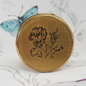 以下が含まれることがあります： 花柄の金色のコンパクト。コンパクトには、表面に刻まれたバラと葉があります。