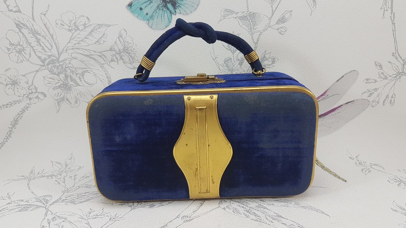 May include: A blue velvet handbag with a gold metal frame and handle. The handbag has a gold metal clasp and a decorative gold metal design on the front.