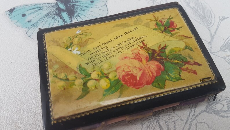 May include: A black, rectangular box with a floral design on the front. The design features a pink rose, white flowers, and green leaves. The box has a gold trim and the text "Think, dear friend, when thou art pondering Orr the past, so sad to thee, With its many scenes and changes, Think of me, Oh! think of me." and "Patent" are printed on the front.
