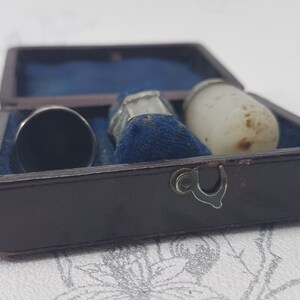 May include: A small, brown leather box with a blue velvet interior. The box contains three thimbles: one silver, one white, and one with a silver top and a clear base.