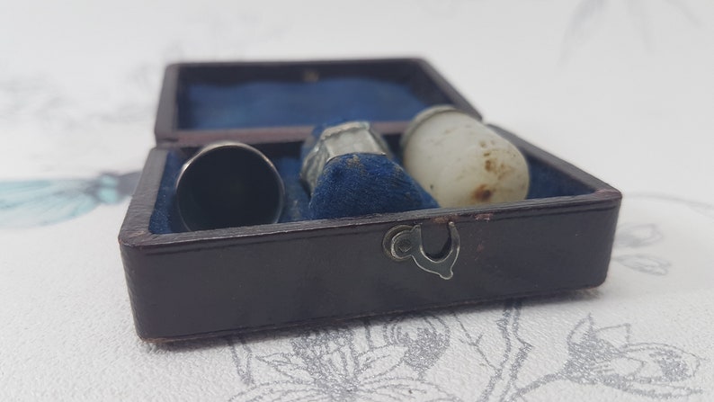May include: A small, brown leather box with a blue velvet interior. The box contains three thimbles: one silver, one white, and one with a silver top and a clear base.