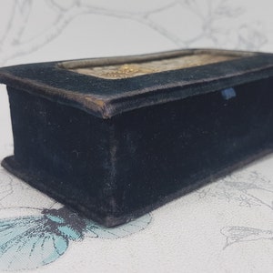 May include: A small, rectangular box covered in black velvet. The box has a glass top and a decorative design on the inside.