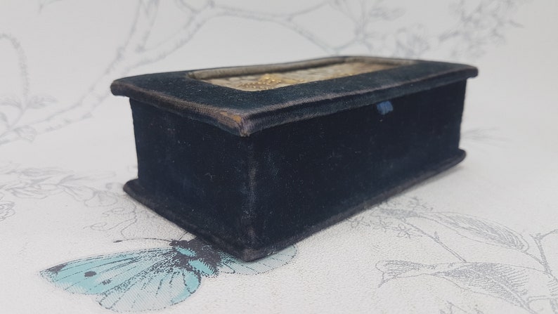 May include: A small, rectangular box covered in black velvet. The box has a glass top and a decorative design on the inside.