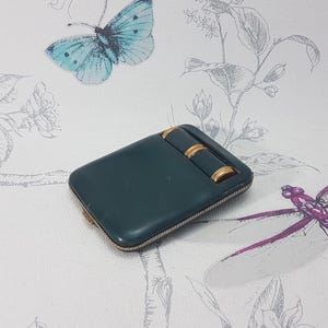 May include: A teal-colored, rectangular case with two gold-colored cylindrical accents. The case has a gold-colored zipper and is set against a floral and butterfly patterned background.