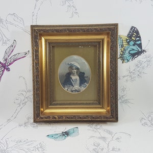 Op de afbeelding: Een gouden lijst met een portret van een vrouw die een blauwe hoed en een blauwe en witte jurk draagt. Het portret is tegen een witte achtergrond met een lichtbruine passe-partout.