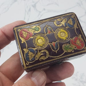 May include: A small, rectangular box with a dark brown leather exterior. The box is decorated with intricate gold and colorful floral designs. The box has a hinged lid and is likely used for storing small items.