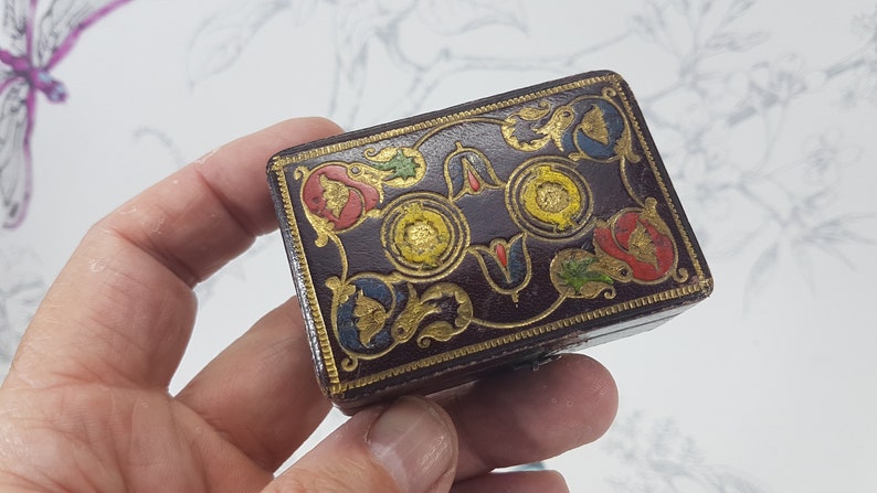 May include: A small, rectangular box with a dark brown leather exterior. The box is decorated with intricate gold and colorful floral designs. The box has a hinged lid and is likely used for storing small items.