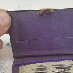 May include: A purple fabric sewing needle case with a flap closure. The case has multiple slots for needles and a small pocket on the inside. The case is open and several needles are visible.
