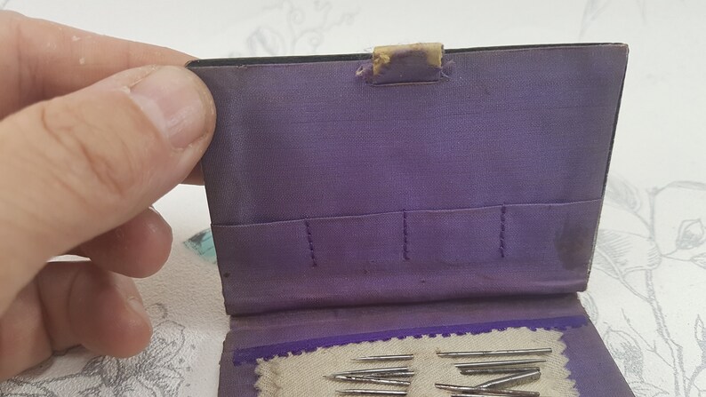 May include: A purple fabric sewing needle case with a flap closure. The case has multiple slots for needles and a small pocket on the inside. The case is open and several needles are visible.