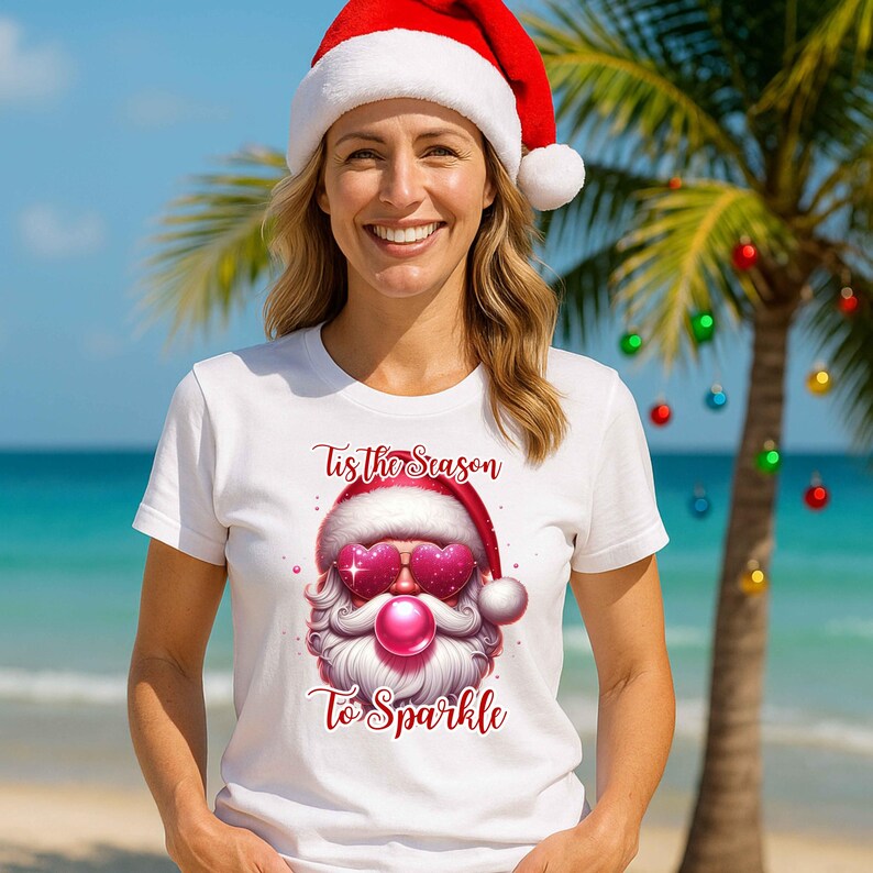 May include: A white t-shirt featuring a Santa Claus graphic with heart-shaped sunglasses and the text "Tis the Season To Sparkle" in red. The Santa wears a red hat with a white pom-pom. The background shows a beach scene.