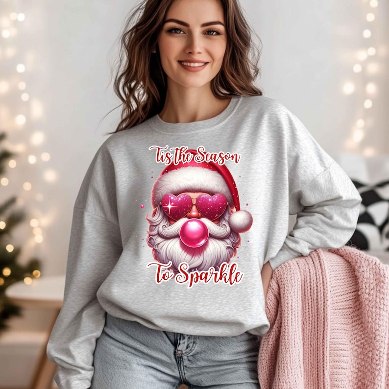 May include: A light gray sweatshirt featuring a Santa Claus graphic with heart-shaped sunglasses and a bubblegum bubble. The text "Tis the Season To Sparkle" is printed above and below the Santa design. The sweatshirt is a festive holiday apparel item.
