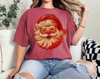 Op de afbeelding: Een bordeauxrood t-shirt met een gedetailleerde illustratie van het gezicht van de kerstman. De kerstman-afbeelding heeft een rode hoed en baard met een glimlachende uitdrukking. Het shirt is een casual stijl met korte mouwen.