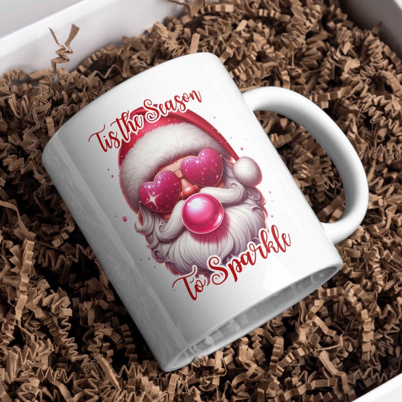May include: White ceramic mug featuring a Santa Claus design with heart-shaped sunglasses and a pink nose. The mug has the text "Tis the Season To Sparkle" in red script. The mug is placed in a box with brown paper.