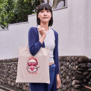 May include: A beige tote bag featuring a Santa Claus graphic with pink sunglasses and the text "Tis the Season To Sparkle." The bag has a simple design with two handles and is being held by a person.