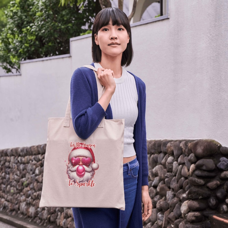 May include: A beige tote bag featuring a Santa Claus graphic with pink sunglasses and the text "Tis the Season To Sparkle." The bag has a simple design with two handles and is being held by a person.
