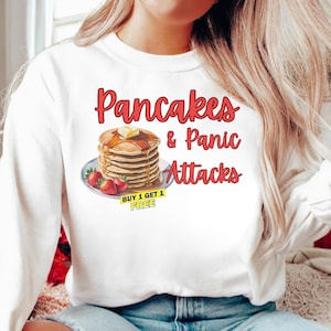 Può includere: Felpa bianca con la scritta "Pancakes & Panic Attacks" in rosso. La grafica presenta una pila di pancake con sciroppo e burro, serviti con fragole. È visibile un'etichetta gialla "Buy 1 Get 1 Free".