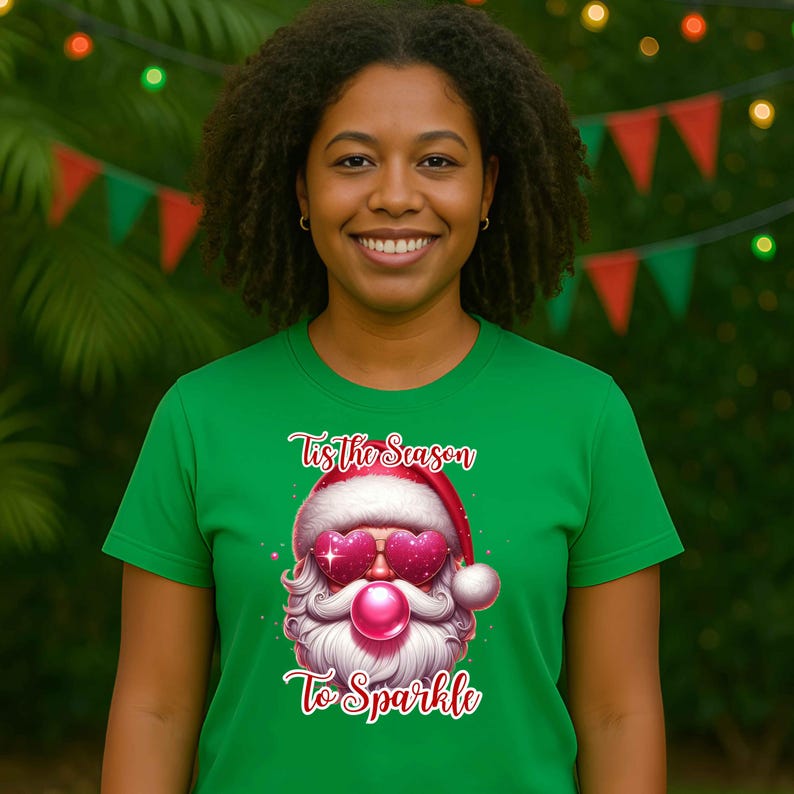 May include: Green t-shirt featuring a Santa Claus graphic with heart-shaped sunglasses and a pink nose. The text "Tis The Season To Sparkle" is written above and below the Santa design. The shirt is worn by a person with dark curly hair.