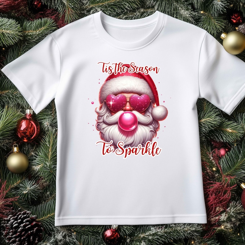 May include: White t-shirt featuring a Santa Claus graphic with heart-shaped sunglasses and a pink bubblegum bubble. The text "Tis the Season To Sparkle" is printed in red. Festive holiday apparel.
