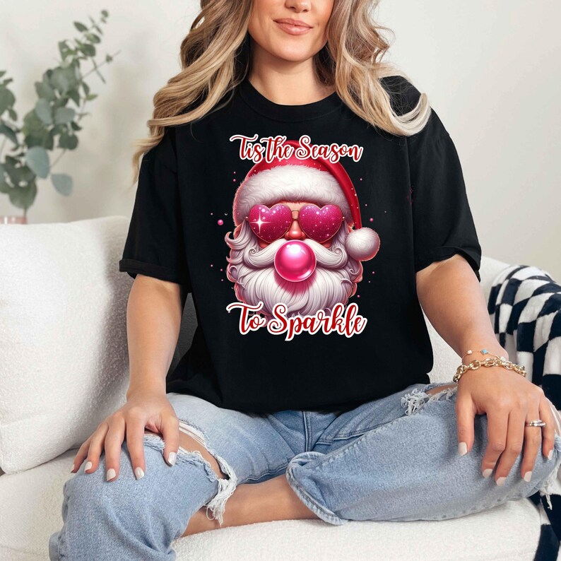 May include: Black t-shirt featuring a Santa Claus graphic with heart-shaped sunglasses and a pink bubblegum bubble. The text on the shirt reads "Tis the Season To Sparkle" in red script. The Santa Claus graphic is wearing a red hat.