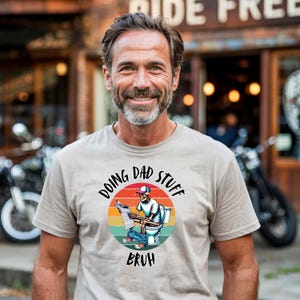 May include: A light gray t-shirt with a retro-style graphic of a skeleton sitting on a toilet and reading a newspaper. The graphic is surrounded by a circle with the text "Doing Dad Stuff" and "Bruh" below it.