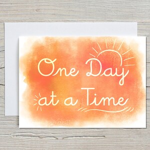 May include: A white greeting card with the phrase "One Day at a Time" in cursive script. The text is in a light orange color, set against a watercolor-style orange and yellow background. A simple sun illustration is above the word "Day".