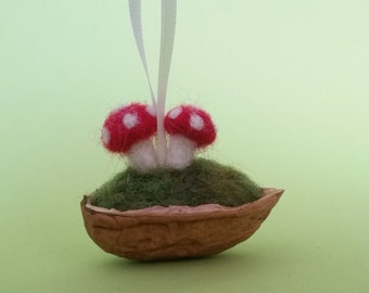 Op de afbeelding: Een grillig ornament met twee rood-witte paddenstoel figuren op groen mos, genesteld in een walnootschil. Een wit lint is bevestigd om op te hangen. De achtergrond is lichtgroen.