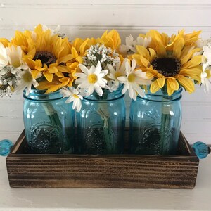 May include: Three blue mason jars filled with yellow sunflowers and white daisies, arranged in a wooden tray with blue glass handles.