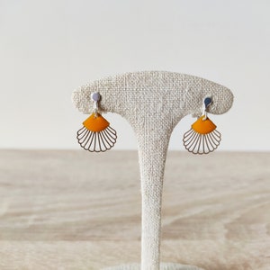 Puede incluir: Un par de pendientes en forma de abanico. Cada pendiente presenta una sección superior de color naranja brillante y una parte inferior plateada calada y festoneada. Los pendientes se muestran sobre un soporte de color neutro.