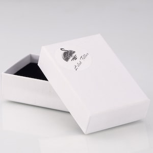 May include: A rectangular white cardboard jewelry box with a black interior lining. The lid features a circular sticker with a black graphic of a flamingo and the text Lilat Filles printed in a cursive script font on the top surface.