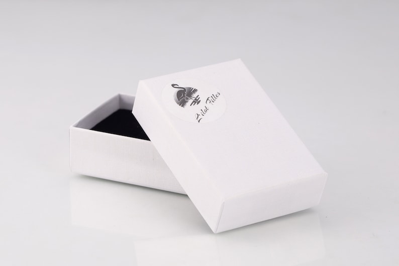 May include: A rectangular white cardboard jewelry box with a black interior lining. The lid features a circular sticker with a black graphic of a flamingo and the text Lilat Filles printed in a cursive script font on the top surface.