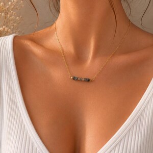 May include: A delicate gold-tone chain necklace featuring a central horizontal bar composed of three small, dark grey, cylindrical stone beads separated by tiny gold-tone metal spacers, resting against a white ribbed fabric garment.
