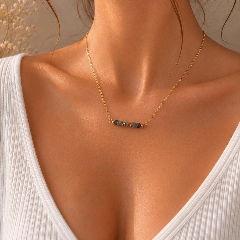 May include: A delicate gold-tone chain necklace featuring a central horizontal bar composed of three small, dark grey, cylindrical stone beads separated by tiny gold-tone metal spacers, resting against a white ribbed fabric garment.