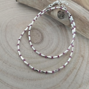 Peut inclure: Un collier en argent délicat à deux rangs, chacun orné de petites perles d'argent facettées et entrecoupées de pierres précieuses rouge-violet. Le collier est présenté sur une surface en bois clair.