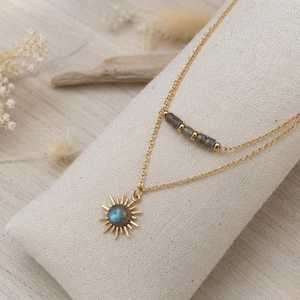 May include: A gold-toned layered necklace featuring two delicate chains. The upper chain displays a row of small grey beads with gold spacers, while the lower chain holds a sunburst pendant centered with a round, iridescent grey gemstone.