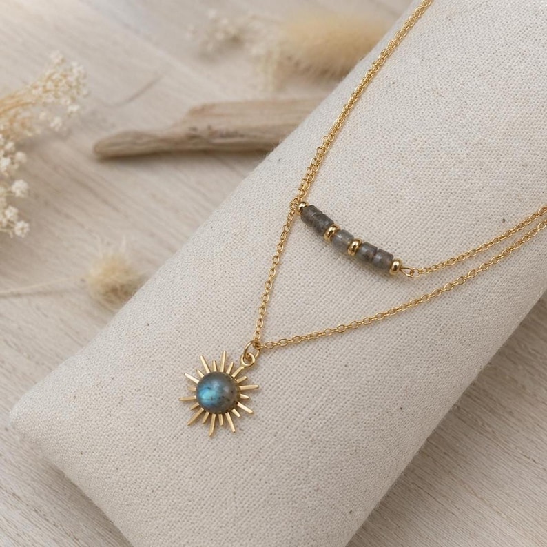 May include: A gold-toned layered necklace featuring two delicate chains. The upper chain displays a row of small grey beads with gold spacers, while the lower chain holds a sunburst pendant centered with a round, iridescent grey gemstone.