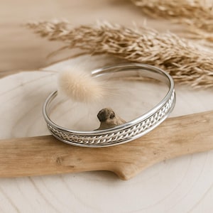 May include: A silver-colored cuff bracelet with a braided design on the front. The bracelet is resting on a piece of driftwood, with a fluffy, light-colored accent and dried plants in the background. The bracelet is a piece of jewelry.