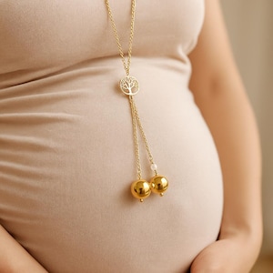 Peut inclure: Un collier doré avec un pendentif arbre de vie et deux breloques sphériques dorées. Le collier est suspendu sur le ventre d'une personne. La personne porte un haut beige clair.