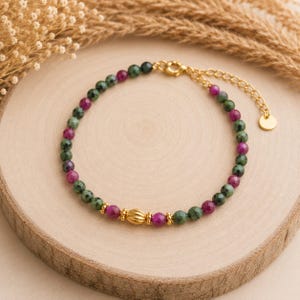 Op de afbeelding: Een kralenarmband met afwisselend gefacetteerde donkergroene en magentakleurige edelsteenkralen, geaccentueerd met een centrale goudkleurige geribbelde kraal en kleine goudkleurige tussenstukjes. Inclusief een goudkleurige verlengketting.