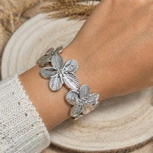 Peut inclure: Un bracelet argenté orné de trois grandes fleurs détaillées. Le bracelet est porté au poignet, avec une manche de pull de couleur claire visible. Les fleurs ont des pétales texturés et sont reliées par une bande.