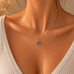 May include: A gold-toned necklace featuring a sunburst pendant with a central circular purple stone. The pendant is suspended from a delicate gold-toned chain that rests against the collarbone. The design includes radiating metal rays.