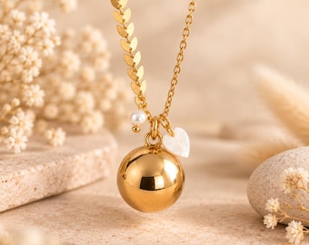 May include: A gold-toned necklace featuring a delicate chain with a leaf-like pattern on one side. The pendant consists of a large, smooth, polished gold sphere, accented by a small white pearl bead and a tiny white heart-shaped charm.