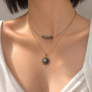 May include: A layered gold-tone chain necklace featuring two strands. The shorter strand includes three small, dark, rectangular beads. The longer strand features a sunburst-shaped pendant with a central, iridescent, round blue-grey stone.