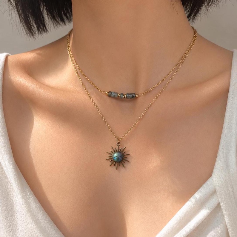 May include: A layered gold-tone chain necklace featuring two strands. The shorter strand includes three small, dark, rectangular beads. The longer strand features a sunburst-shaped pendant with a central, iridescent, round blue-grey stone.