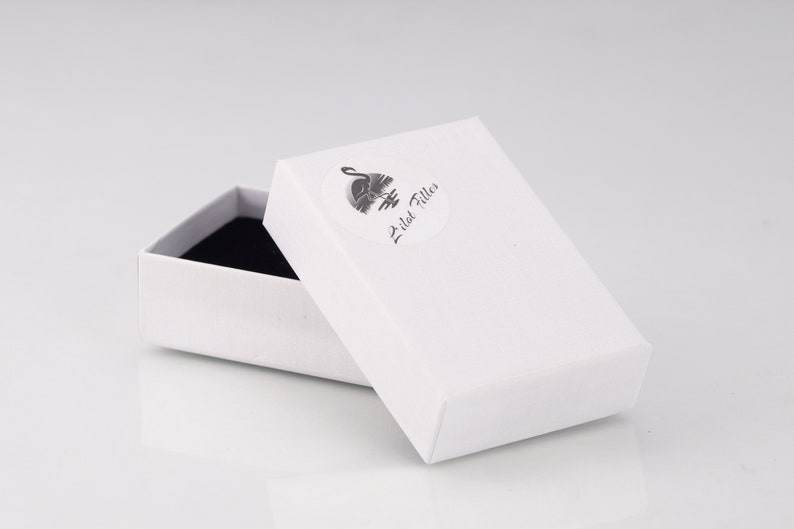 May include: A white rectangular box with a partially opened lid, revealing a black interior. A circular sticker with a swan logo and the text "Lilat Filles" is on the lid. The box is on a white surface.